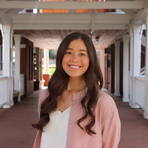 Alyson Sato, standing in the middle of a hallway. She is wearing a pink cardigan and white blouse, with hair down over her shoulders and smiling at the camera. The hallway has red tile and white walls, with wooden frames around the windows and doorways.