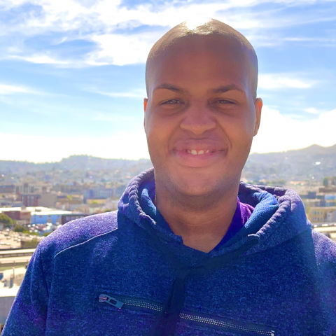 Antoine thomas is facing the camera, smiling. He is wearing a blue/violet hoodie. Behind him is a light blue sky with cirrus clouds whisping over the horizon, and below the sky is a suburban setting with hills in the distance.