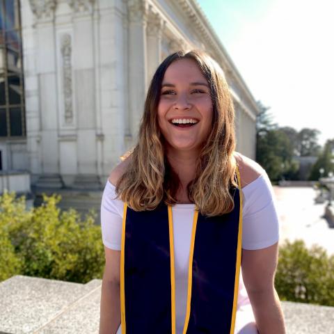 Autumn Kleinman wearing a white dress, smiling. She is wearin gthe UC Berkeley graduation stoll with one of the UC Berkeley buildings behind her.