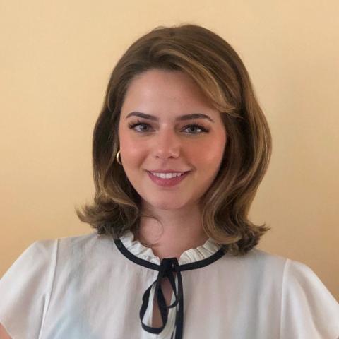 Vjola Jorgji is facing the camera directly, smiling. She is wearing a white blouse with a black ribbon tied around the base of her neck. Behind her is a cream colored wall.