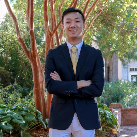 Michael Zhu is standing in the center of the frame, smiling at the camera, with his right arm crossed over his left. He is wearing a black blazer over a grey shirt, yellow tie, and white pants. Behind him is a tree and shrubs.