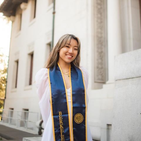 Vivian is standing in the middle of the frame, smiling at the camera. She is wearing her Berkeley graduation stole over her white dress in front of a white building. 