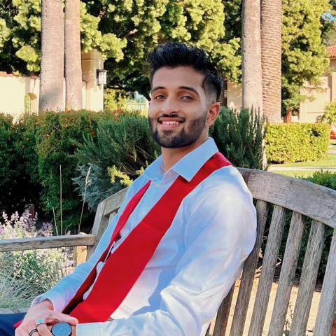 Abhijit is pictured sitting on a bench, turned slightly to the right while turning his head to the left to smile directly at the camera. He is wearing a light dress shirt with a red graduation stole. 