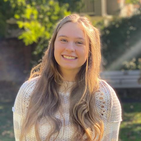 Hannah is standing in the middle of the frame, smiling at the camera, her hair laid over her shoulders. She is wearing a white sweater. 