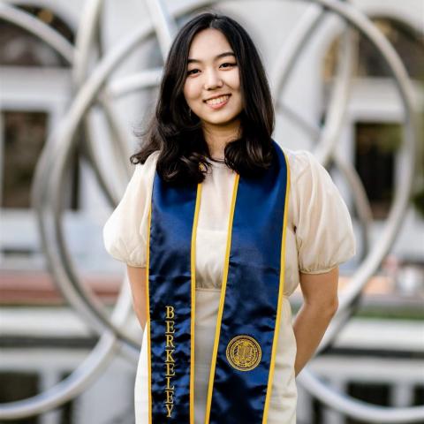 Sydney Yoon is standing in the middle of the frame, smiling at the camera. She is wearing a light sundress with a blue and gold graduation stole with the word Berkeley over her right shoulder and the University of California insignia over her left. 