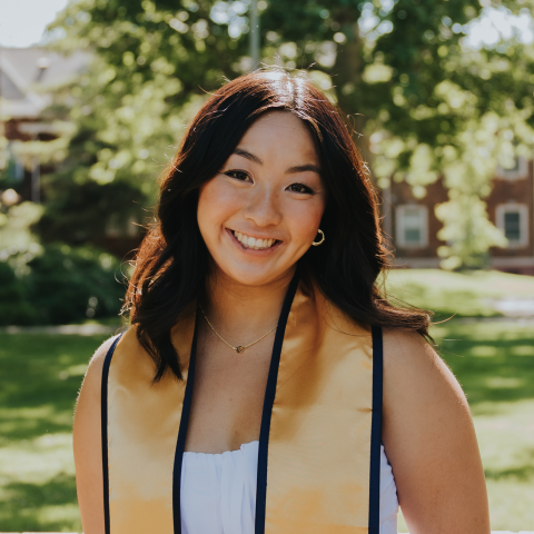 Samantha is standing in the middle of the frame, facing the camera and smiling. She is wearing a white dress. She has a blue and gold UC Davis graduation stole draped over her shoulders. 