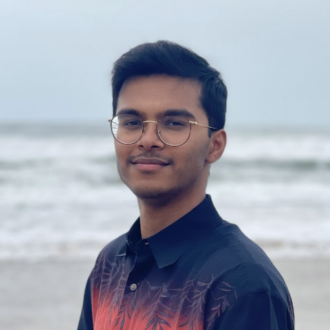 Shreyas is standing in the center of the frame, facing the camera and smiling. Behind him is the ocean with an overcast sky.. 