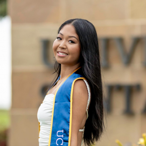 Jazlene is facing the left of the frame, her head is turned towards the front of the camera, smiling. She is wearing a white dress and an over-the-shoulder graduation stole for UC Santa Barbara.