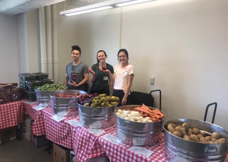 NHC SF AmeriCorps Members (From left to right, Wesley, Julia, Kristin) standing at the HealthRIGHT 360 Food Pharmacy table, with bowls of fruits and vegetables set up on top of the table. 