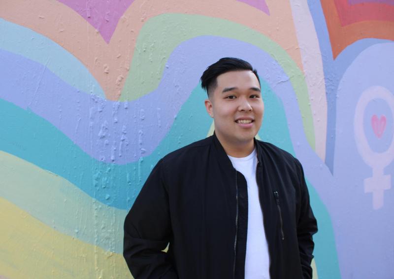 NHC SF member Antony Nguyen, smiling, wearing a black jacket over a white t-shirt. The background is a mural of overlapping, colorful painted hearts, next to a drawn symbol.