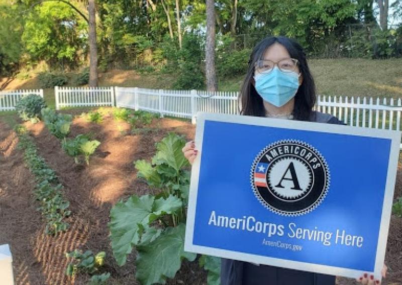 Jennifer standing in a garden and holding a sign that reads "AmeriCorps Serving Here"