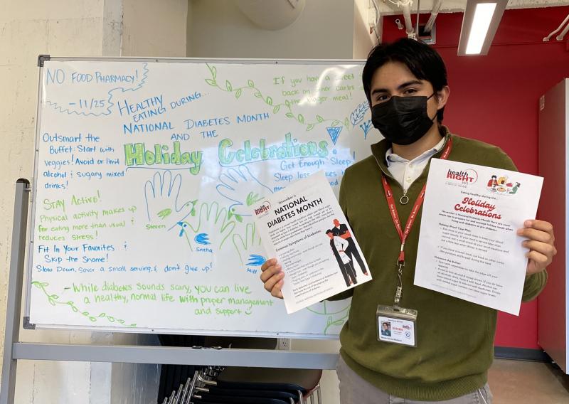 Steven is seen here facing the camera, holding up two educational material flyers regarding nutrition and diabetes care awareness. To his right is a white board with writing at the center about healthy eating during the holiday months for patients with diabetes.