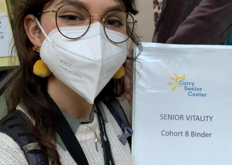 Sarah is facing the camera. She is holding a binder that reads "Curry Senior Center; Senior Vitality; Cohort 8 Binder". 