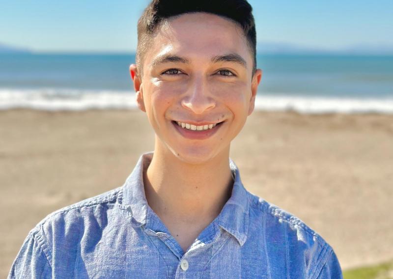 Cameron is standing in the middle of the frame, smiling at the camera. Behind him is a beach, with the sand followed by the sea in the distance.