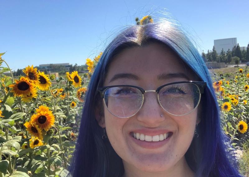 Megan is facing the camera smiling. Behind her is a field of sunflowers and a clear, blue sky.