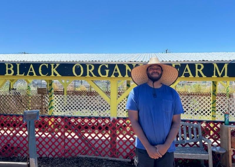 Faheem Carter stands in the center-right of the frame. Behind him is a farm shed with the sign "Black Organic Farm" painted onto the wooden overhang. 