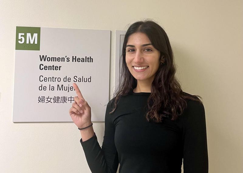 Anisha is seen in the center of the frame, facing the camera and smiling. She is pointing at a sign that reads "5M: Women's Health Center." The sign is also translated into Spanish and Chinese.