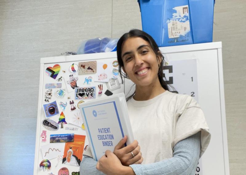 Anjalie is standing in the center of the frame, holding a binder. The cover of the binder reads "Patient Education Binder."