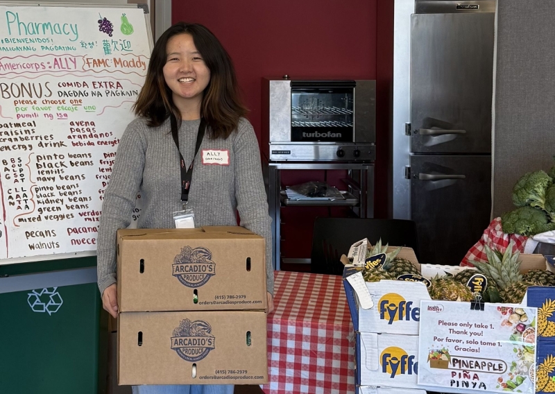 Ally is standing to the left of the frame holding two boxes of produce. Behind her is a white board explaining what is in each box, and to the right of her are stacked boxes filled with a variety of vegetables.