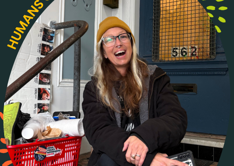A frame with the text "Humans of NHC" and "Mary, Program Director, Homeless Youth Alliance" circles the photo of a woman sitting on the top of a staircase leading to a front door. She is facing the camera and smiling. Next to her right arm is a grocery basket filled with materials, including compostable cups and tote bags, among other resources.