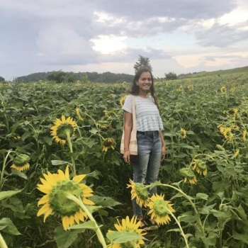 NHC PGH member Roosha smilling, standing in a sunflower field on an overcast day