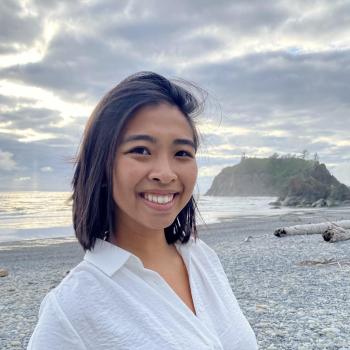 Emily Pham, facing the camera and smiling. Emily is wearing a white blouse. The sky is cloudy with some rays of sunlight breaking through over the grey-ish sand, ocean. There is an island in the distance