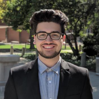 Jack Dokhanchi facing the camera directly and smiling. Jack is wearing a black suit jacket with a blue dress shirt buttoned up to the top button, and glasses sit squarely on his face. There is a tree in the background, and a courtyard with concrete floors, a grassy area, and a red wall in the distance.