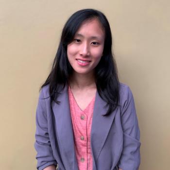 Mindy is facing the camera smiling, wearing a lavender dress coat over a pink blouse. Behind her is a cream-colored, blank wall.