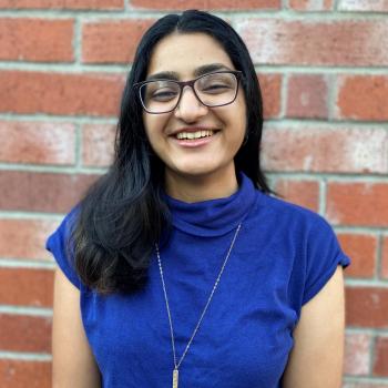 Saadhana is facing the camera, smiling. Their hair is draped over their right shoulder. They are wearing a blue short sleeve turtleneck, with a necklace hanging over their chest. Behind them is a brick wall with both fully red and faded red bricks.