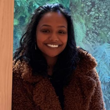 Sneha is facing the camera directly and smiling. She is behind a cream colored pillar and she is wearing a coat. Behind her is a window facing out to a wall with plants hanging over the threshold.