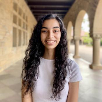 Anita stands in the middle of the photo frame, smiling at the camera. Behind her is Stanford Main Quad arcade with the columns to the right. 