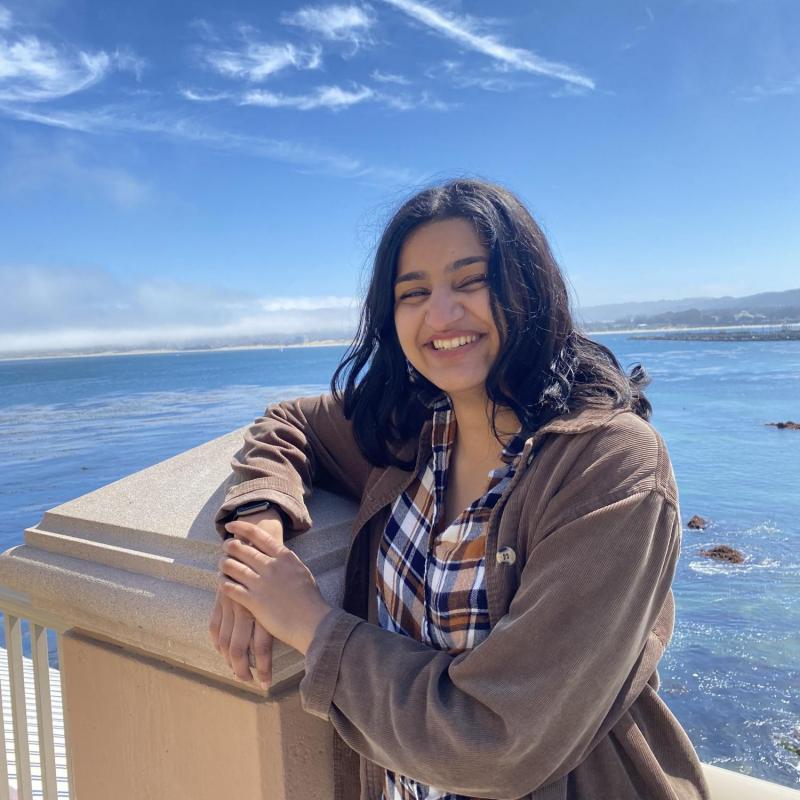 Saadhana is seen facing the camera, smiling. They are wearing a beige coat over a plaid-patterned buttoned shirt loosely tucked into light-wash jeans. They are leaning on a post. Behind them is the ocean with some light clouds in the sky. 