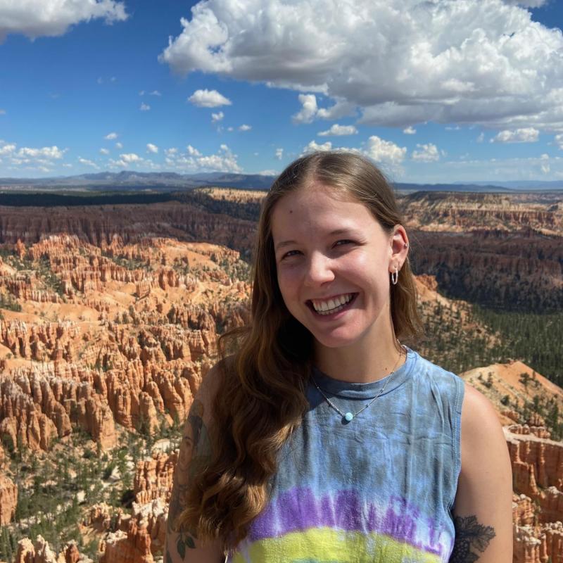 Grace Lemke is standing to the middle right of the frame, smiling at the camera. She is wearing a tie-dyed tank top. Behind her is a canyon with mountains in the far background. 