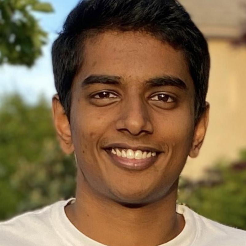 Sai is centered in the frame, facing the camera and smiling. He is wearing a white shirt.
