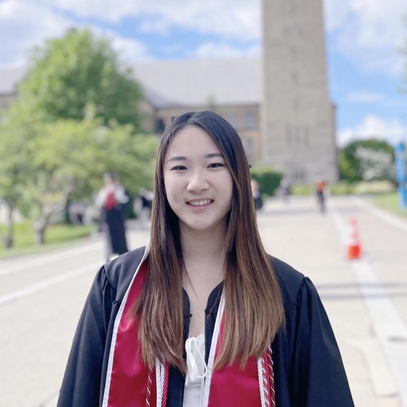 Joelle is standing in the middle of the frame. She is wearing a red stole over graduation regalia and a white dress. Behind her is a building that has been blurred out in the distance. 