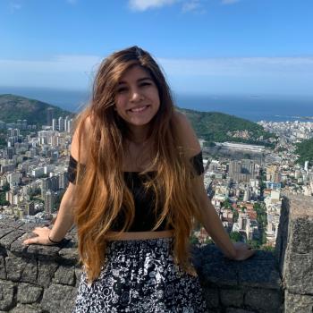 Paola is standing in the middle of the frame, leaned against a low stone railing and smiling at the camera. Behind her is a view of a city, rolling hills, and the ocean in the distance.
