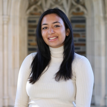 Rachel Mundaden is seen here facing the camera, smiling. She is wearing a white tutrtleneck sweater. Behind her is a gothic archway. 