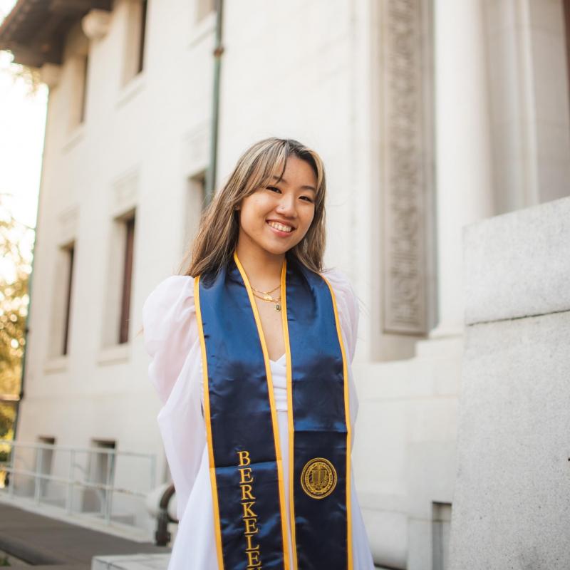 Vivian is standing in the middle of the frame, smiling at the camera. She is wearing her Berkeley graduation stole over her white dress in front of a white building. 