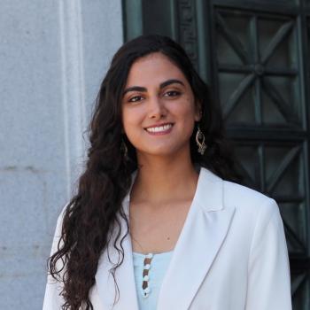 Alya is standing in the middle of the frame, facing the camera and smiling. She is wearing a white shirt under a white blazer.