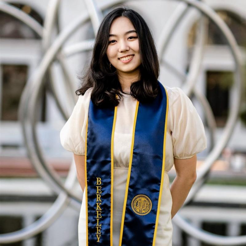 Sydney is standing in the middle of the frame, smiling at the camera. She is wearing a light sundress with a blue and gold graduation stole with the word Berkeley over her right shoulder and the University of California insignia over her left. 