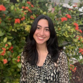 Laura is standing in the middle of the frame, facing the camera and smiling. Behind her is a bush of flowers. 