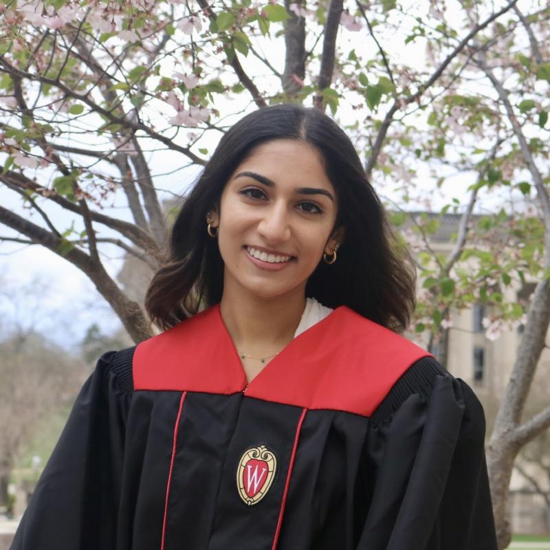 Anisha is standing in the middle of the frame, facing the camera and smiling. She is wearing black graduation regalia with red accents. Behind her is a tree with sparse foliage.