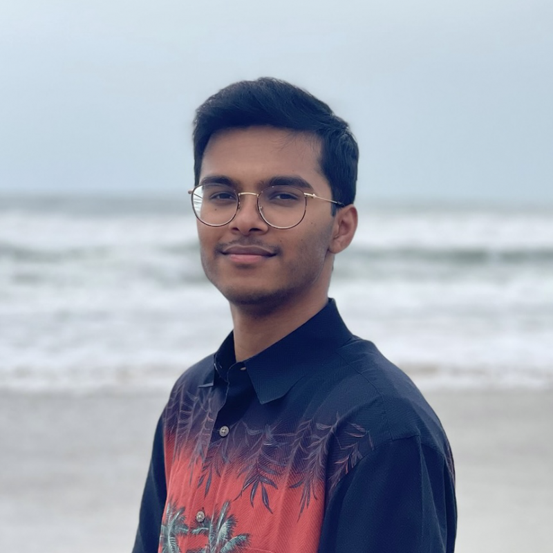 Shreyas Raj is standing in the center of the frame, facing the camera and smiling. Behind him is the ocean with an overcast sky.. 