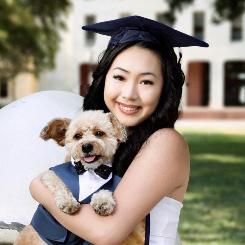 Renee is centered in the middle of the frame, facing the camera and smiling. She is carrying a fluffy, white dog. The dog is dressed in a tuxedo and bowtie. Renee is wearing a white dress and a graduation cap. 