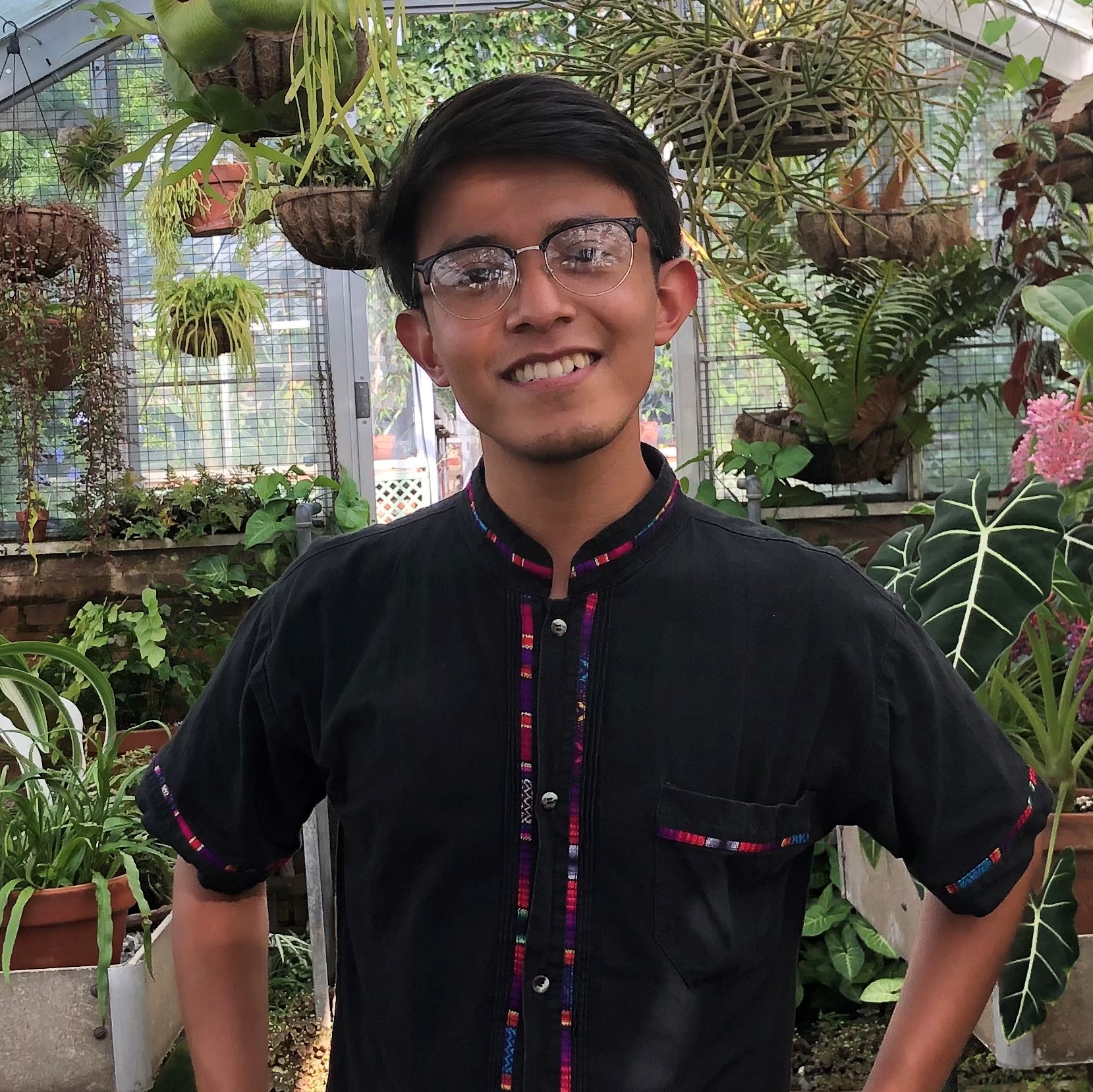 Alex stands in the center of the frame, smiling at the camera. His arms are placed on his hips. Behind him is a room filled with a variety of different plants.