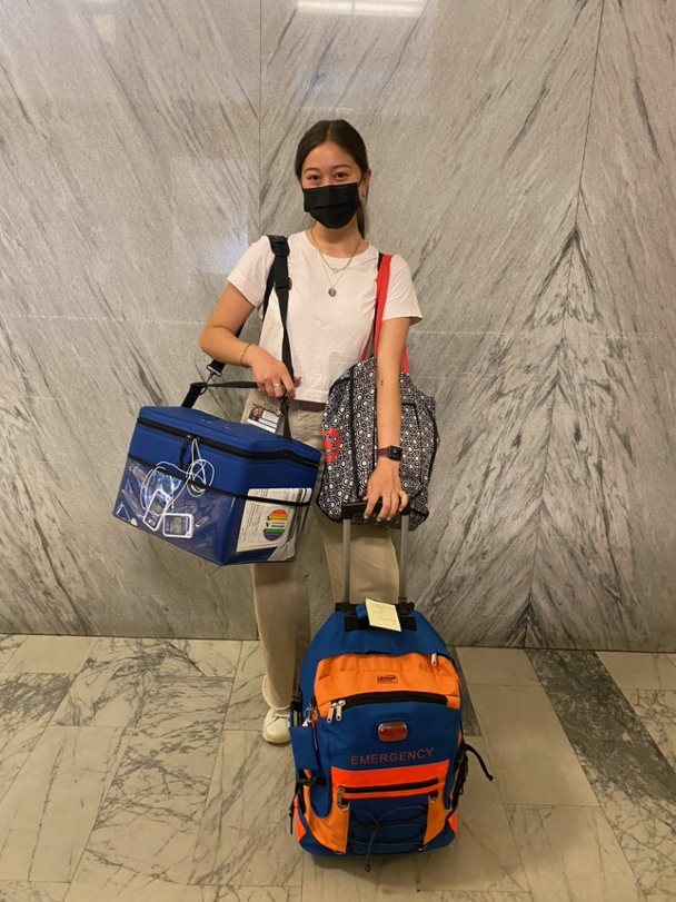 Aly is standing in the middle of the photo. She is holding one bag over her right shoulder, another bag over her left shoulder, as well as holding the handle of a third rolling backpack. The third backpack has the word "Emergency" printed across the face of the bag.