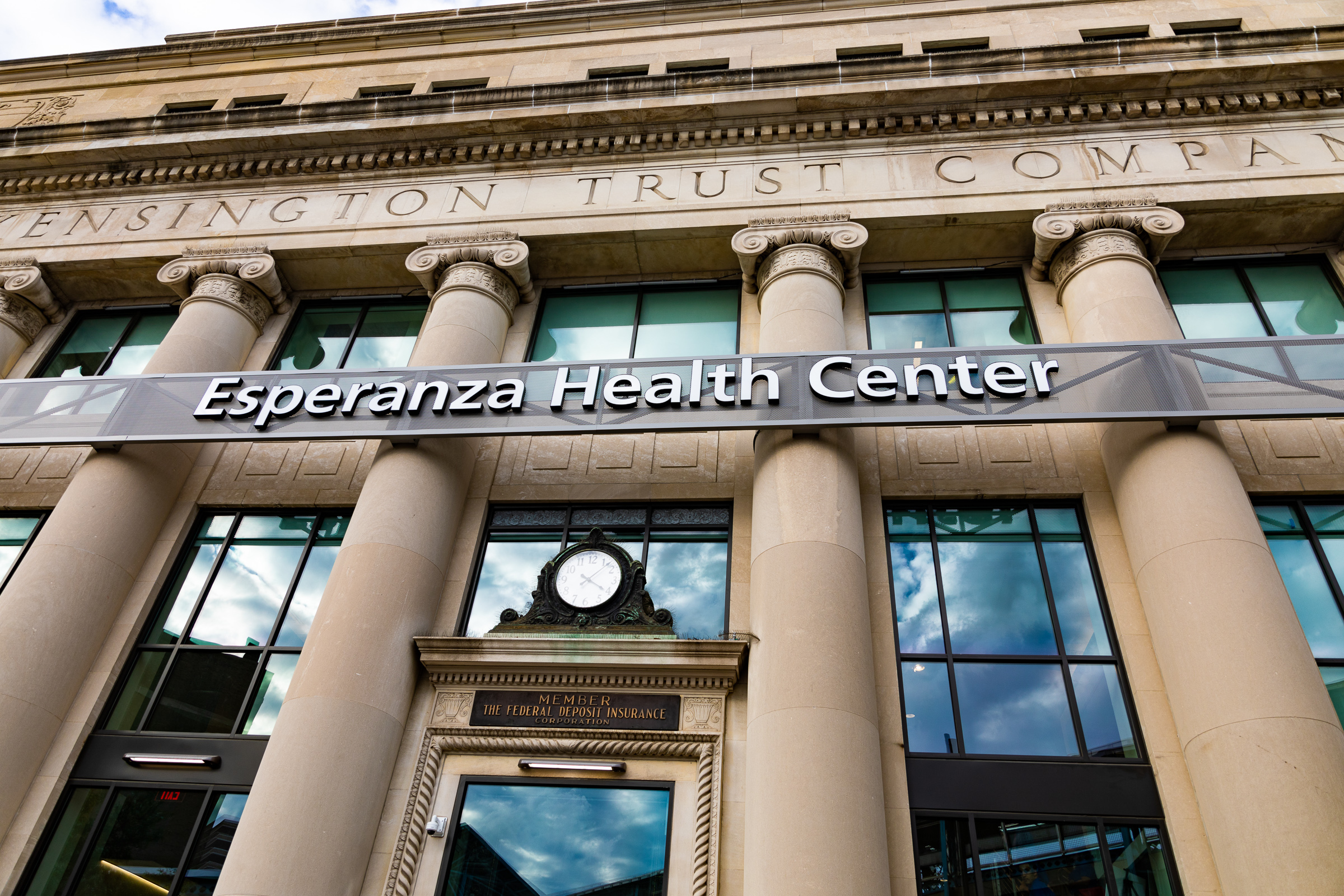 Looking up at a building with tan columns. There is an ornate clock over the front door and above that a sign that says "Esperanza Health Center"