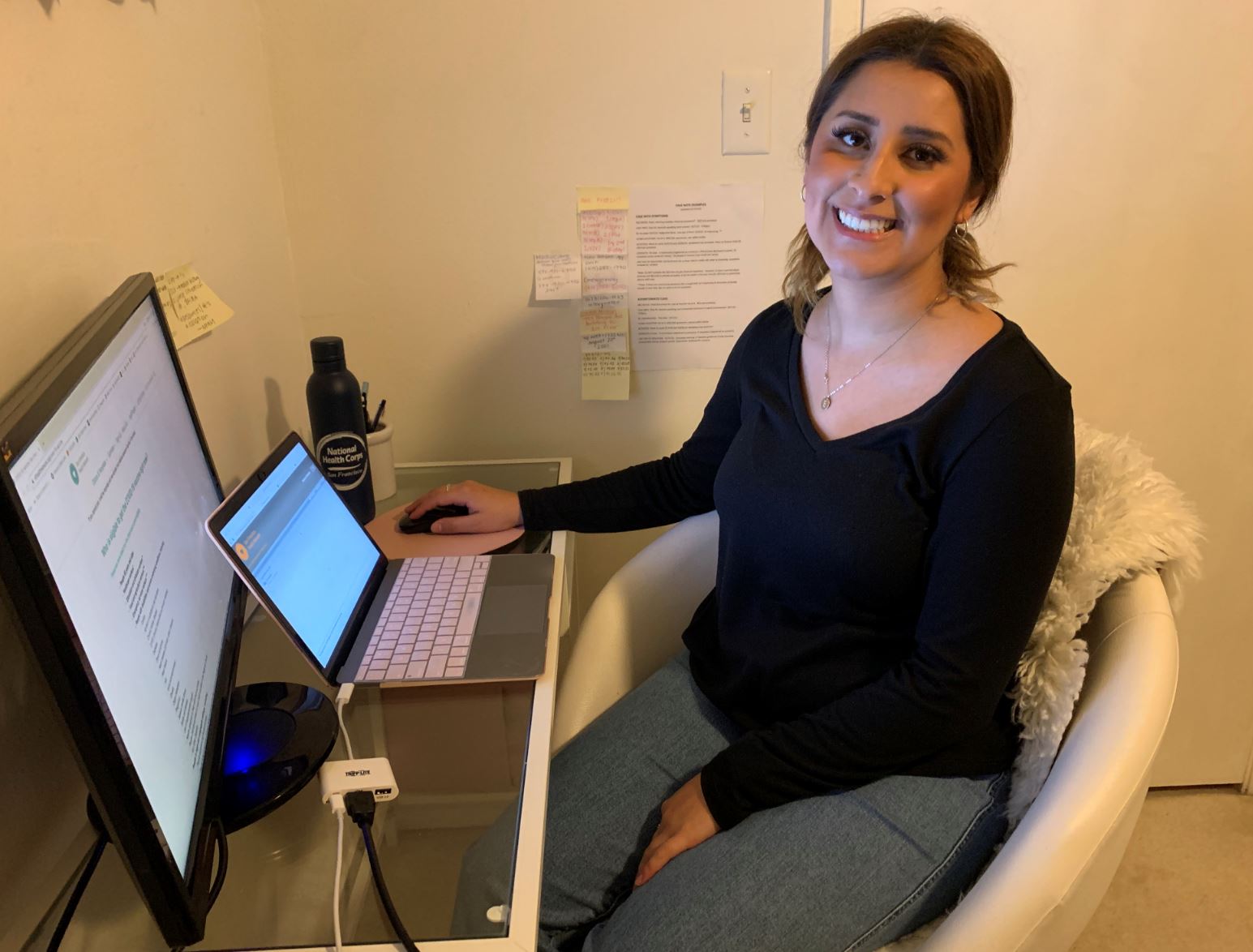 Evelyn De Arcos, NHC SF AmeriCorps Member, sitting in a white comfy chair in front of her laptop and monitor screen, which are both set atop of her desk, while serving from home. 