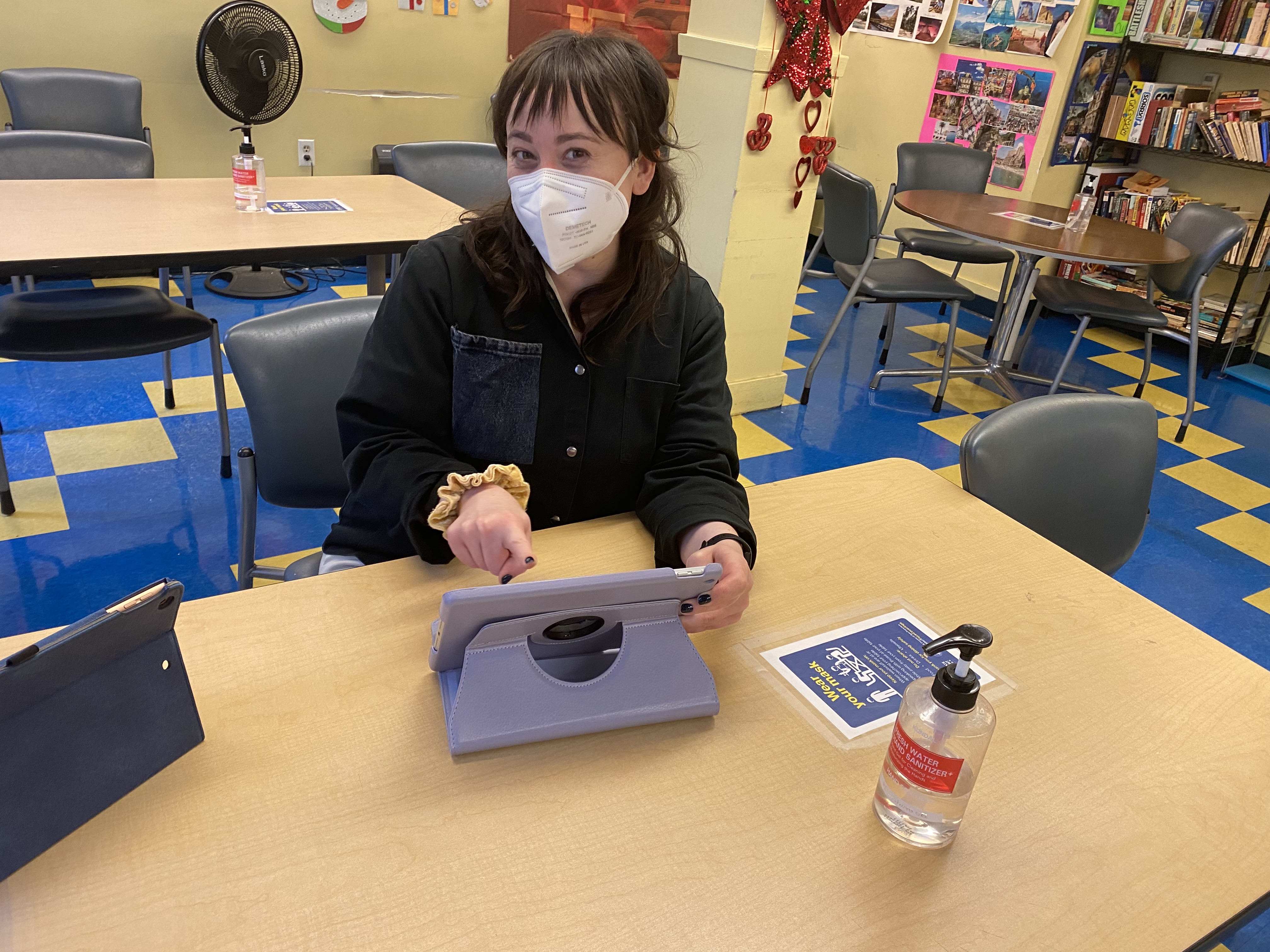 Hannah is seen facing the camera while pointing at an iPad, guiding an older adult (off-screen) during an iPad tech support session.