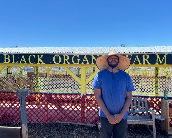 Faheem Carter stands in the center-right of the frame. Behind him is a farm shed with the sign "Black Organic Farm" painted onto the wooden overhang. 